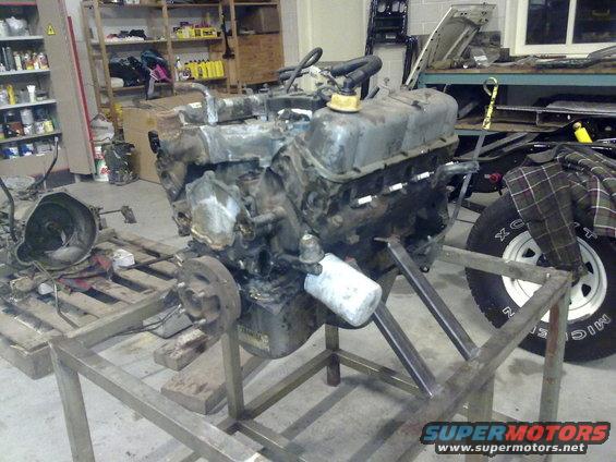Engine on a metal stand in a workshop, with a transmission and tire visible in the background