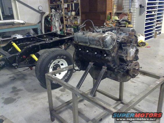 Vehicle frame with rear axle and suspension, next to a disassembled engine on a stand. Workshop setting