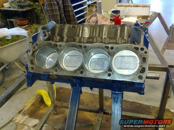 Engine block with four pistons visible, mounted on a stand. Yellow hose nearby