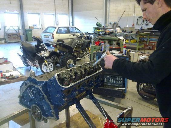 Engine block on stand with person working on it, scooter, ATV, and car in background