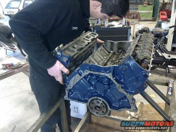 Man holding a blue engine block with the cylinder heads removed, showing internal components