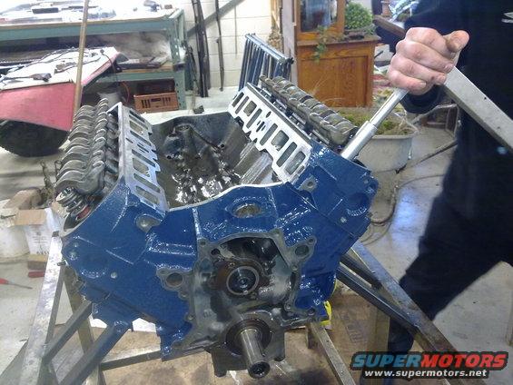 Blue engine block with valve covers removed, on a metal stand. A person's arm is visible operating a tool