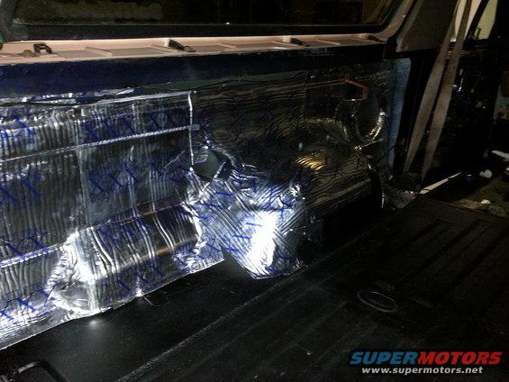 Interior of a vehicle with silver and blue sound dampening material covering the rear side panel and black carpeted floor