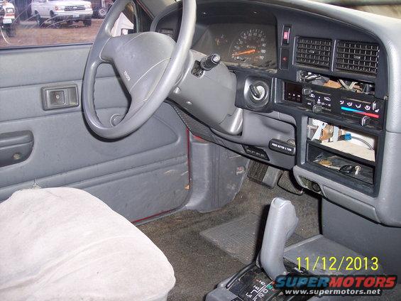 Interior of a red Toyota pickup truck, showing the dashboard, steering wheel, and gear shifter. A white pickup truck is visible outside the driver's side window