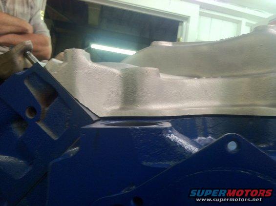 Close-up of a blue engine block with a white intake manifold on top. A person is visible in the background