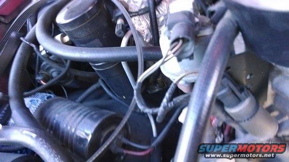 Engine compartment with hoses, wires, and a black cylindrical component. Watermark reads "Supermotors.net"