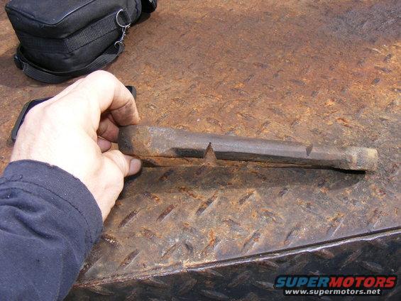Hand holding a rusty metal tool with a pointed end on a diamond plate surface. A black bag is in the background