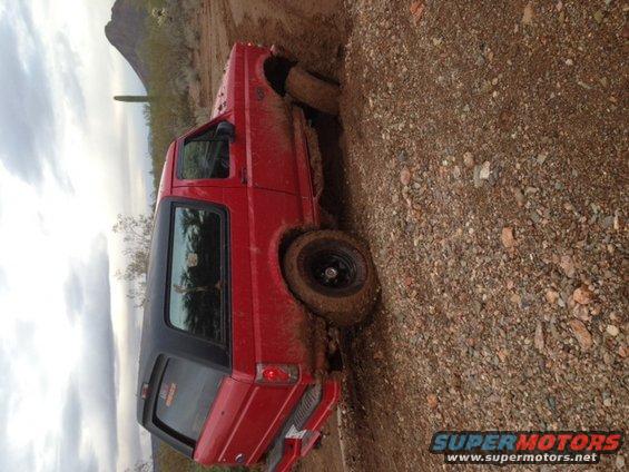 Red SUV stuck on a muddy hill, front wheel deep in dirt. Desert landscape with cactus in background