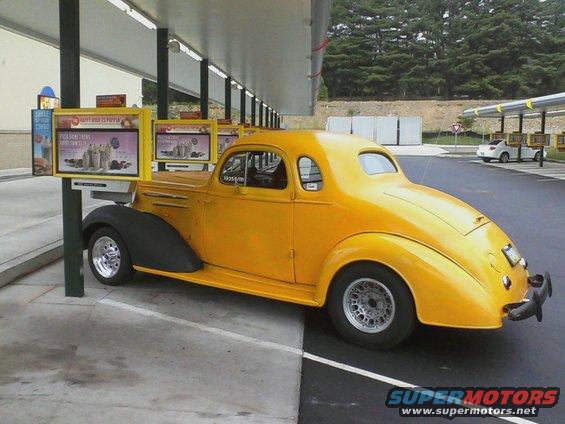 Yellow 1935 Chevrolet coupe hot rod parked at a drive-in restaurant with menu boards. Another white car is visible in the background