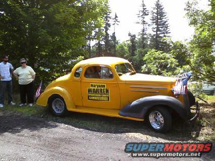 Yellow 1936 Chevrolet coupe with "MARYANNE WARREN INDIANA COUNTY COMMISSIONER" on door. Two people stand near front