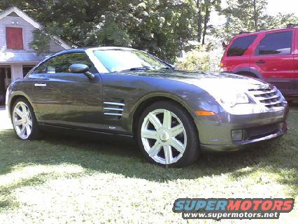 Gray Chrysler Crossfire parked on grass, front-left quarter view. Red SUV behind