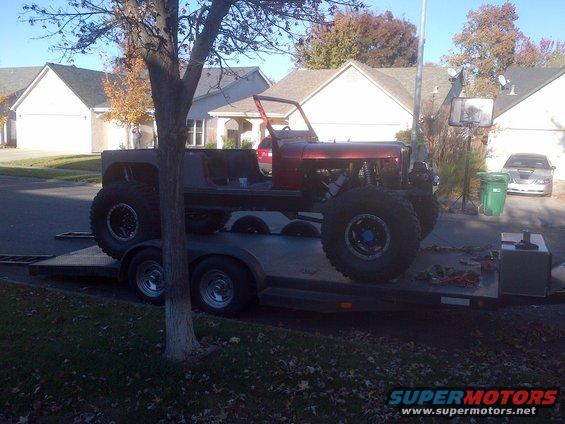 Custom off-road vehicle with large tires on a trailer. Red body, exposed engine components, and roll cage visible