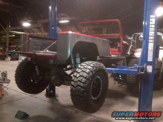 Red Jeep CJ-series body on a lift, with custom flatbed and large off-road tires. The vehicle is in a workshop