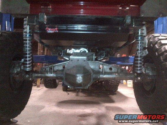 Rear axle assembly with large coilover shocks and massive tires. A red body panel is visible above