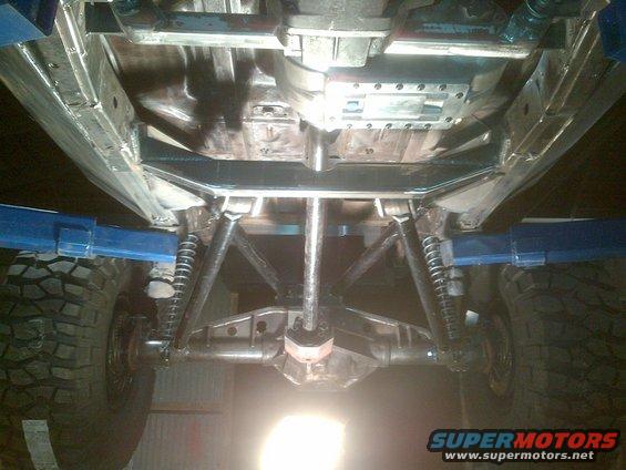 Underside of a vehicle on a lift, showing suspension and drivetrain components, with large off-road tires