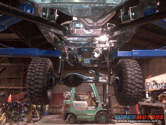 Underside of a Jeep chassis on a lift, with large off-road tires and a forklift below