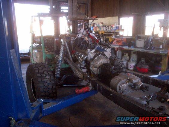 Jeep CJ frame with engine and transmission installed, on a lift. Forklift in background