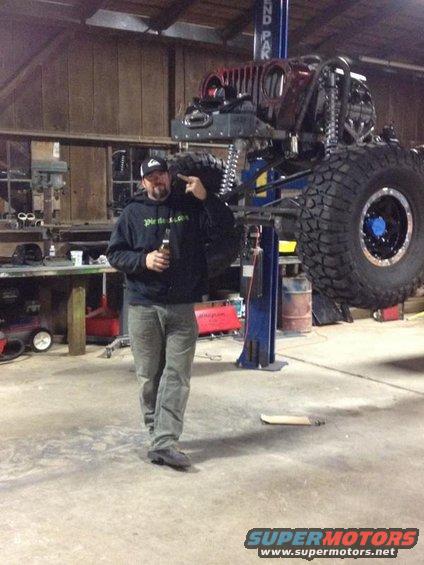 Man in garage points at heavily modified Jeep lifted in the air. It has large tires and custom suspension