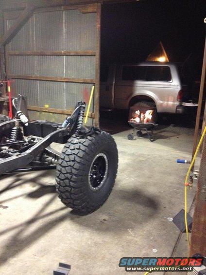 off-road vehicle chassis with large tire in foreground, pickup truck in background with fire pit