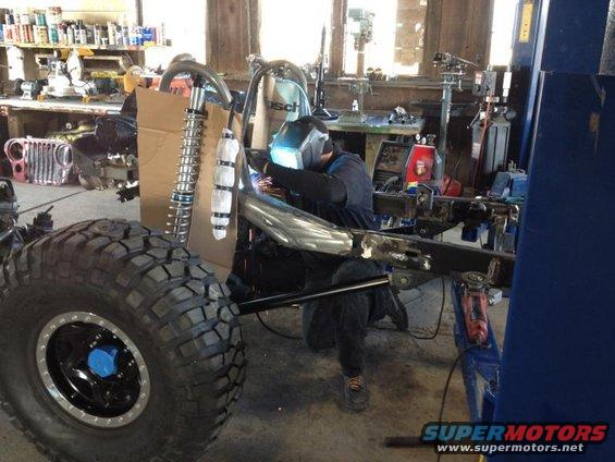 Man welding Jeep CJ frame in workshop, large tire in foreground