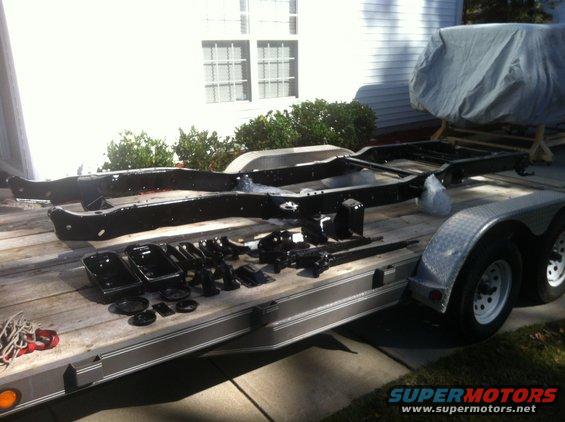 Vehicle frame and various suspension components laid out on a trailer. A tarp-covered object is visible behind the frame