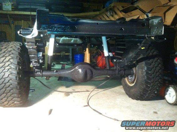 Vehicle frame with rear axle, leaf springs, and shocks visible. Large tires are mounted