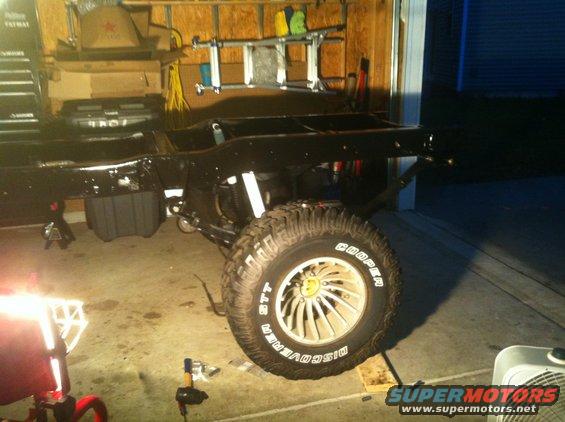 Black vehicle frame and suspension with large off-road tire on a jack stand in a garage
