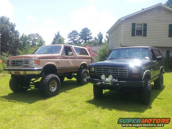 Two lifted Ford Broncos parked on a lawn in front of a house. One is tan and brown, the other is black