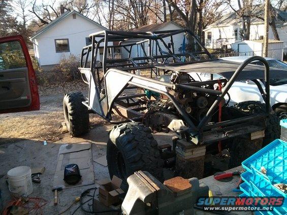 Custom off-road buggy frame with engine and large tires, partially assembled outdoors. Red door visible on left