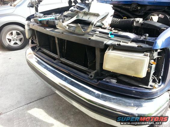 Front of a blue truck with the grille and headlight removed, showing the engine bay. Another car is parked next to it