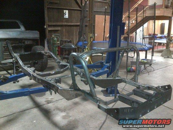 Jeep CJ frame with custom roll cage welded on, sitting on a lift in a garage. A partial vehicle body is visible to the left