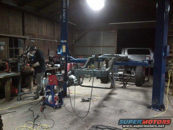 Jeep CJ frame on a lift with a welder working nearby and a silver pickup truck in the background