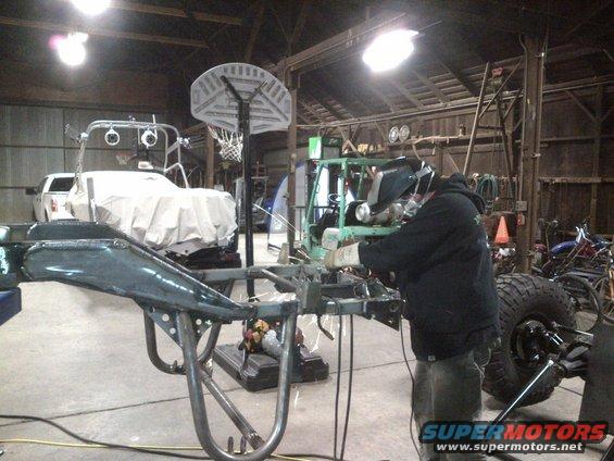 Welder working on a vehicle frame in a garage. Another vehicle is covered in the background