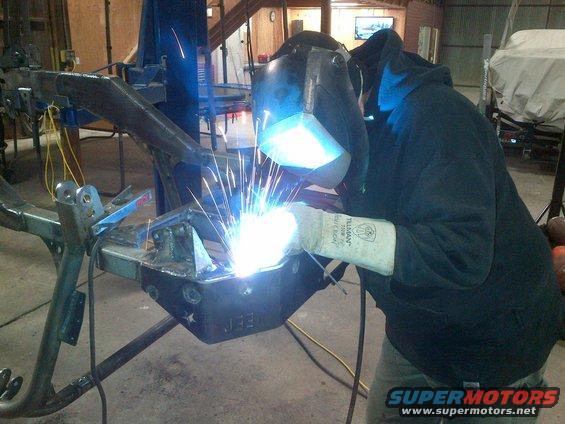 Jeep CJ frame section with "JEE" stamped on it, being welded by a person in a mask and gloves