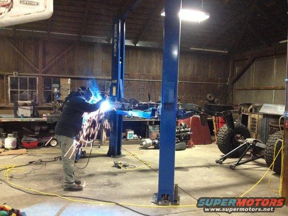 Jeep CJ frame and large tires on a lift in a workshop with a person welding