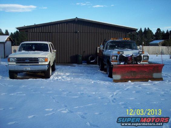 Two white Ford trucks parked in snow, one with a snow plow