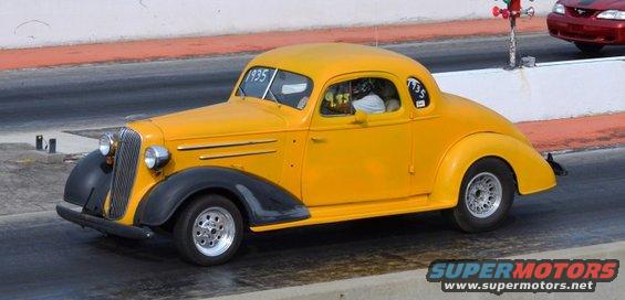 Yellow 1935 Chevrolet coupe hot rod with black fenders and chrome wheels on a race track