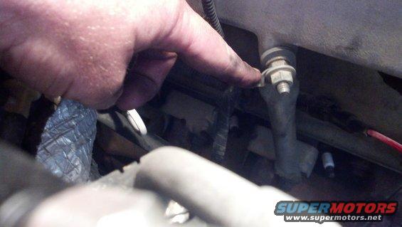 Hand pointing to a bolt on a metal engine component with hoses and wires nearby