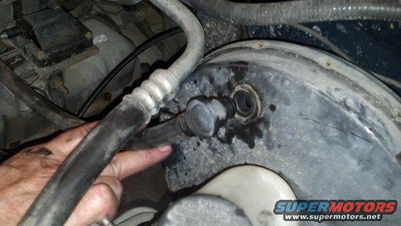 Close-up of a brake booster with a vacuum line disconnected. A hand is pointing to the connection point