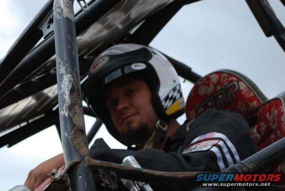 Man wearing a white Bell helmet with checkered flag pattern, seated in a vehicle with a roll cage. A red seat with floral pattern is visible