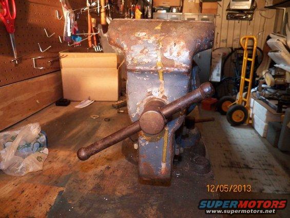 Rusty bench vise with a large handle, set on a wooden workbench. Tools hang on a pegboard behind it