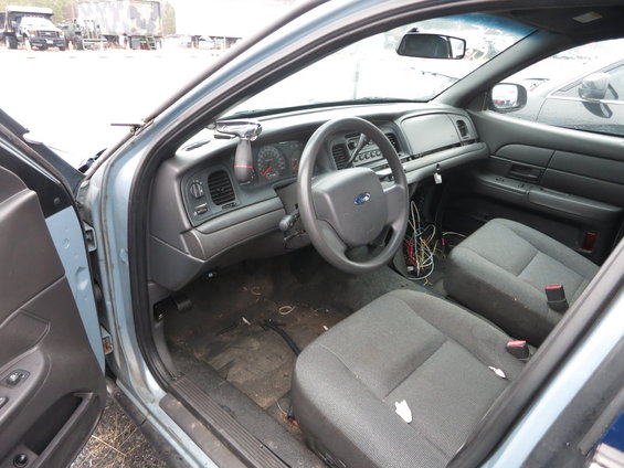 Interior view of a blue police car, showing the dashboard, steering wheel, and front seats. Wires are visible near the center console