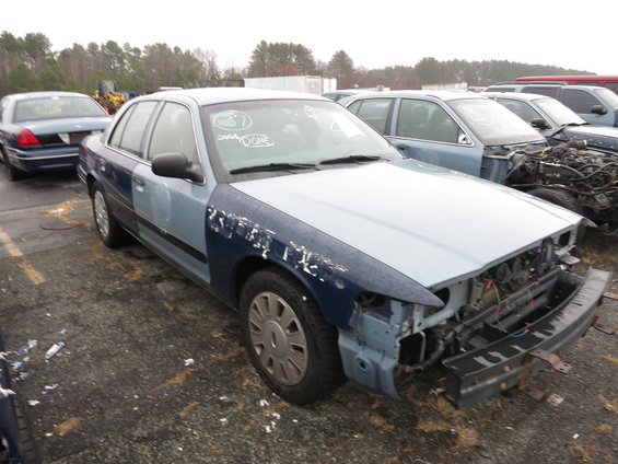 Blue and light blue sedan, missing front bumper and grille, in a junkyard