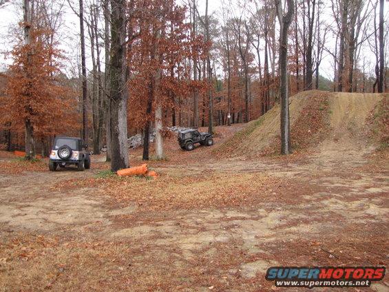 Two off-road vehicles parked in a wooded area with fallen leaves and a dirt ramp
