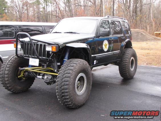 Modified Jeep Grand Cherokee with oversized off-road tires, custom front bumper, and blue suspension components. Another vehicle is partially visible to the left
