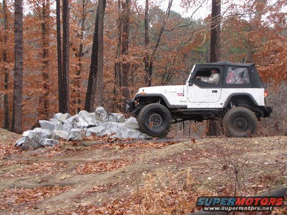 White Jeep Wrangler with a black soft top driving over rocks on a wooded trail