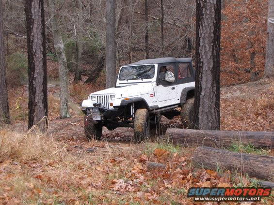 White Jeep Wrangler with soft top navigating a wooded trail, tires on uneven ground