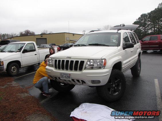 White Jeep Grand Cherokee with lifted suspension, two people working on front bumper. White pickup truck parked nearby
