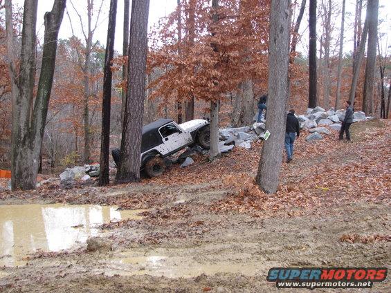 White Jeep Wrangler navigating a muddy, wooded terrain with rocks