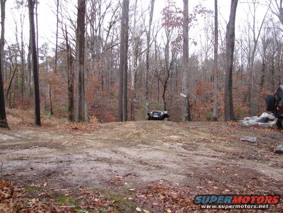 Jeep Wrangler on dirt hill, another vehicle visible on right. Woods in background